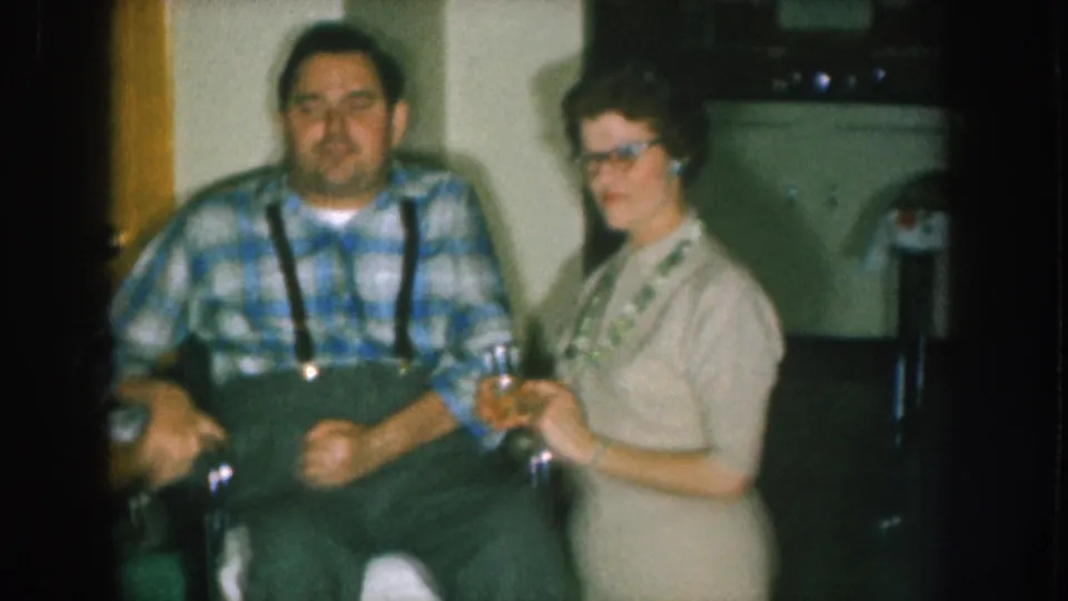 Final frame of 8mm sequence circa 1957 Nebraska; man in wheelchair in blue plaid shirt and suspenders beside woman wearing glasses, both appear to hold drinking glasses, interior setting.