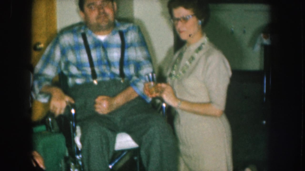 1957 Nebraska home movie — man in wheelchair wearing blue plaid shirt and suspenders; woman in cat-eye glasses and beige sheath dress holds a glass beside him in a domestic interior.