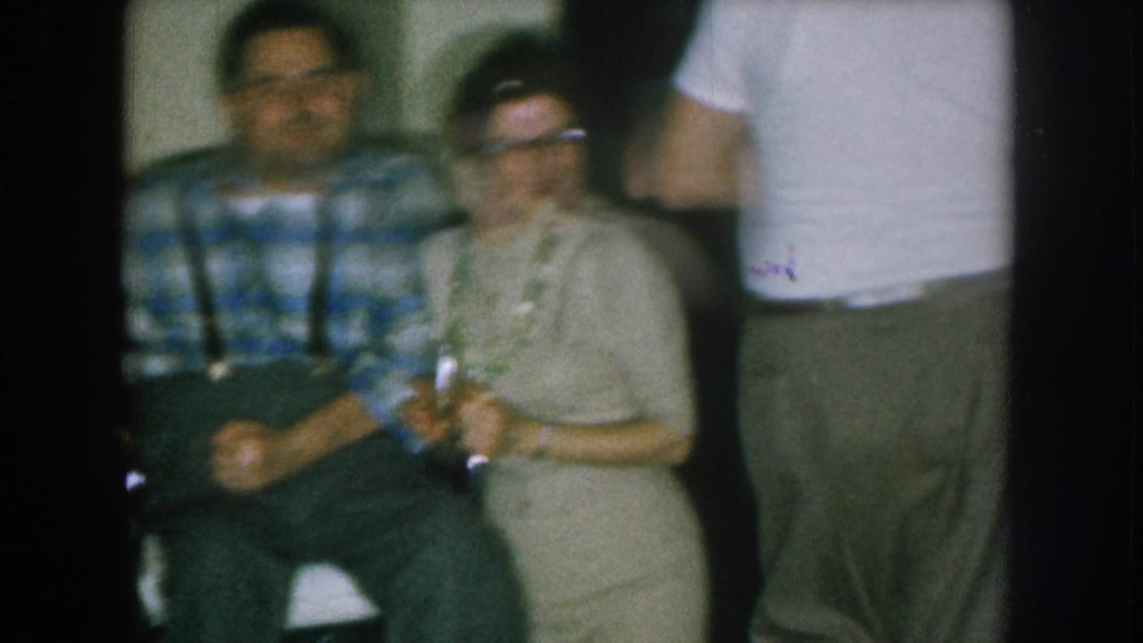 Seated man in plaid shirt and suspenders with woman in glasses; third person in white shirt partially visible at right edge of frame.