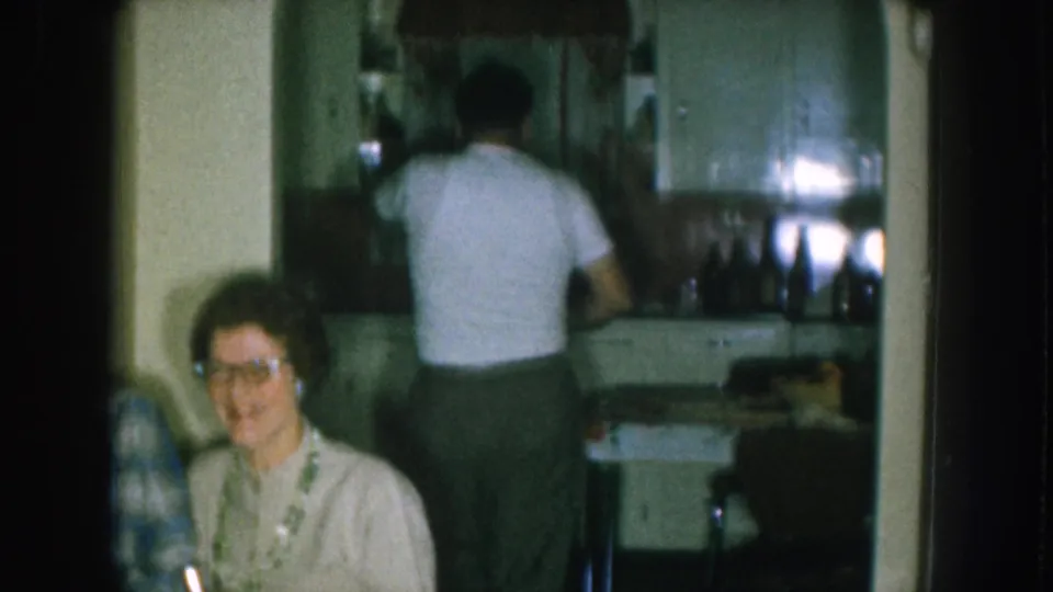 1957 Nebraska home movie — smiling woman with cat-eye glasses and necklace seated in foreground; man in white shirt at a kitchen counter lined with bottles in the background.