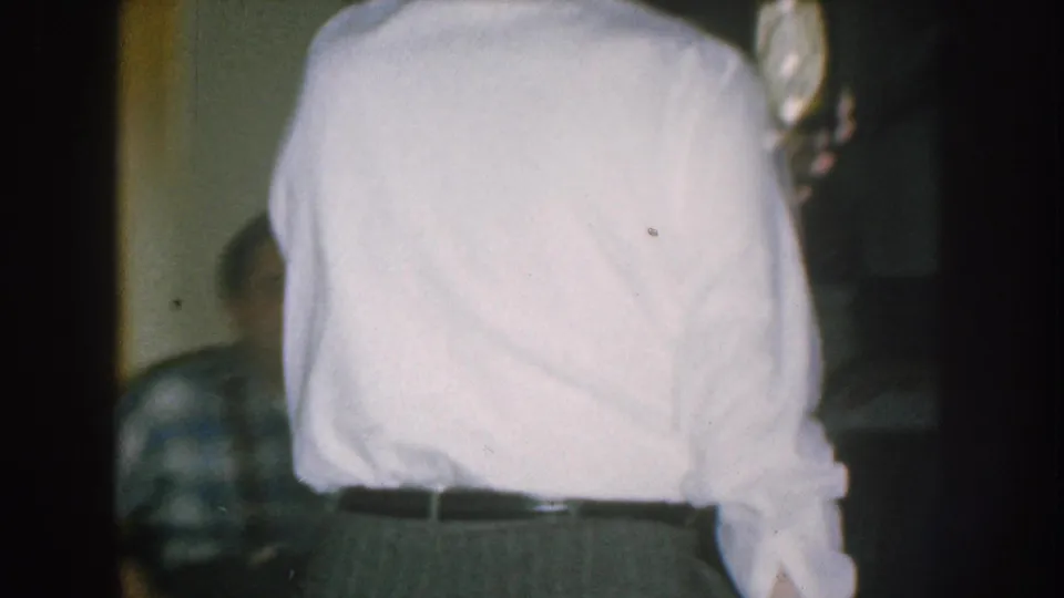 1957 Nebraska home movie — tight rear shot of man in white shirt; second figure in plaid shirt partially visible at lower right.