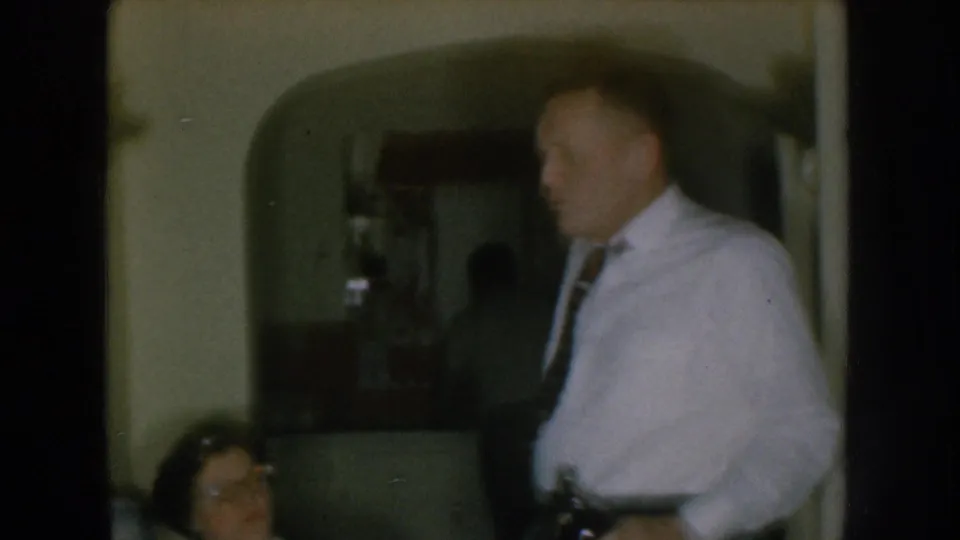 Man in white shirt and tie crossing a domestic room, 1957 Nebraska 8mm home movie; arched doorway and partially visible seated woman at left.