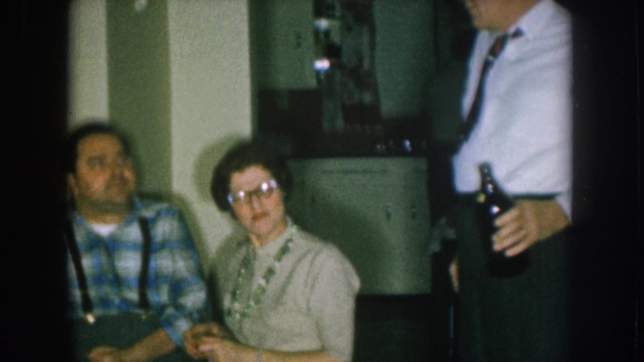 Man in blue plaid shirt and suspenders seated beside a woman with cat-eye glasses, with a man holding a dark bottle at right; Nebraska 1957 8mm home movie.