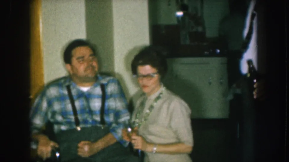 Man in blue plaid shirt with suspenders and a woman with cat-eye glasses in a domestic interior, Nebraska 1957 8mm; a bottle-holding figure is partially cropped at right.