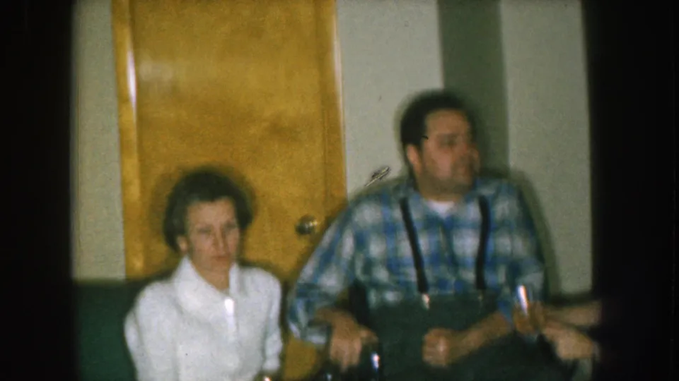 Interior domestic scene with a prominent wooden door, a woman in white blouse, and a man in blue plaid shirt with suspenders; Nebraska 1957 8mm home movie.