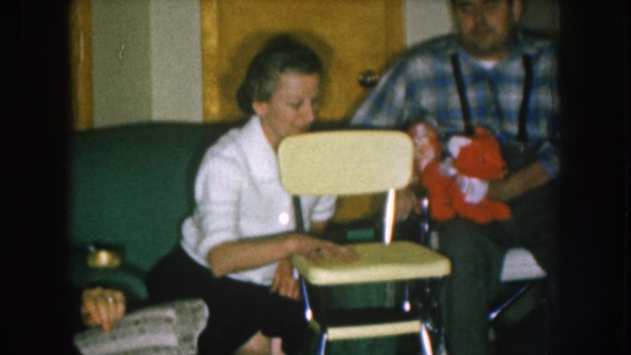 1957 Nebraska home movie: woman in white cardigan and man in blue plaid shirt with suspenders displaying a small yellow child's chair and red stuffed toy indoors.