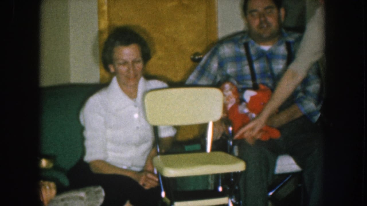 Couple flanking a small yellow child's chair with an off-camera arm reaching in from the right; man holds a red stuffed toy.