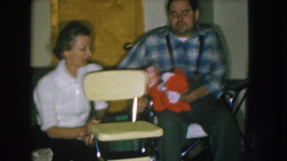 1957 Nebraska home movie: wider framing of couple with yellow child's chair and red stuffed toy, green upholstered furniture and a wooden door visible in the background.