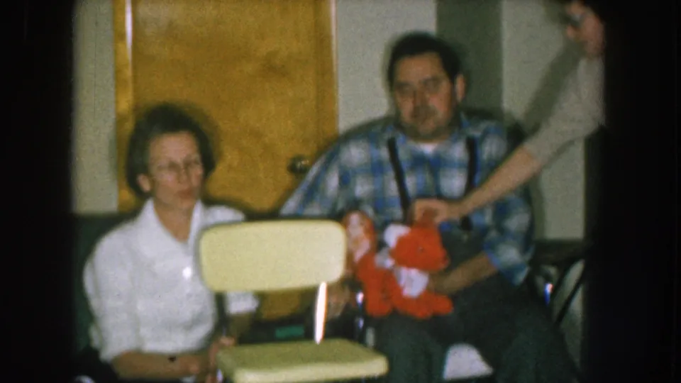 1957 Nebraska home movie: man in plaid flannel and suspenders holding red stuffed toy beside woman with yellow child's chair, an off-camera arm visible at frame right.