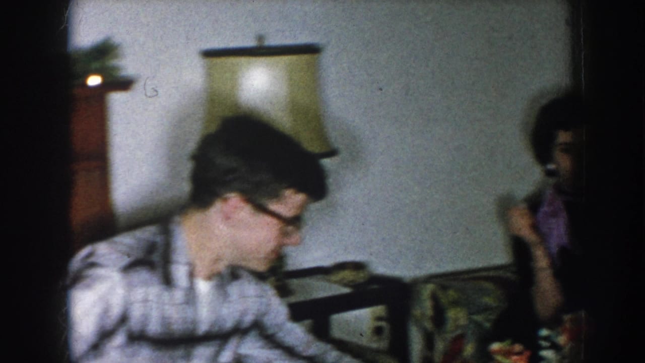 Young man in plaid shirt and black-rimmed glasses in a domestic living room, Nebraska USA, circa 1957; woman in dark dress visible in background.