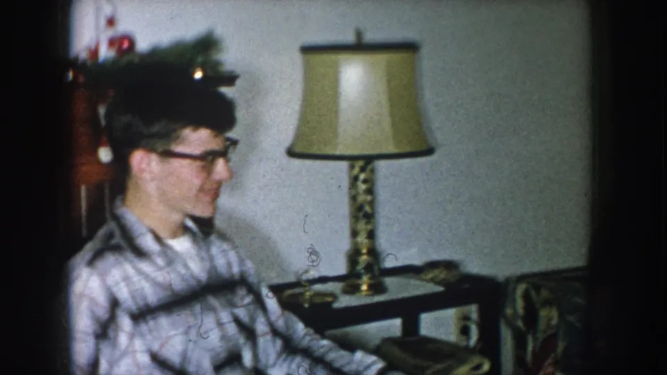 Young man in plaid shirt and heavy black-rimmed glasses smiling in a living room, Nebraska USA, circa 1957; brass table lamp and holiday greenery behind him.