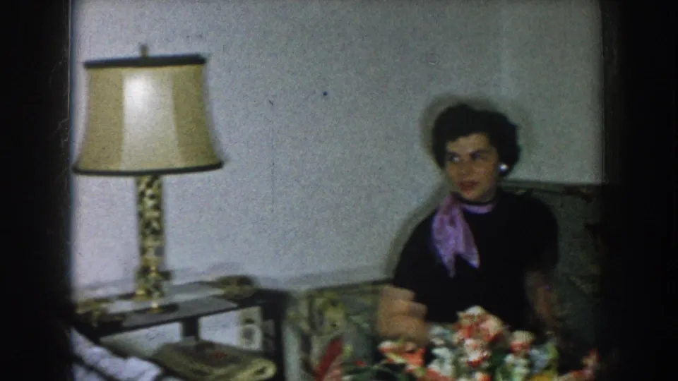 Woman in dark dress and purple scarf seated on floral upholstered couch in a domestic living room, Nebraska USA, circa 1957; table lamp and holiday items visible.