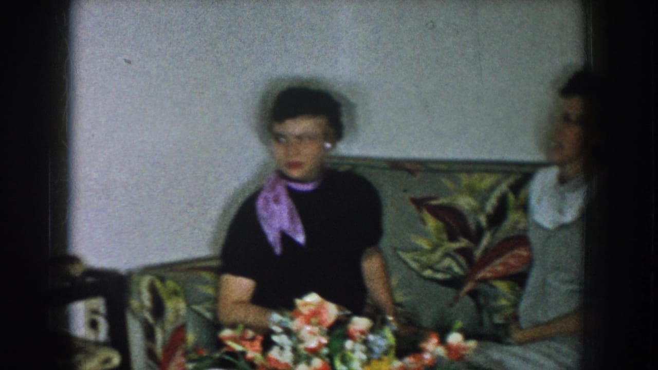 Two women in late-1950s daywear on a floral-upholstered sofa in a domestic living room; a colorful flower bouquet sits on a surface in the foreground.