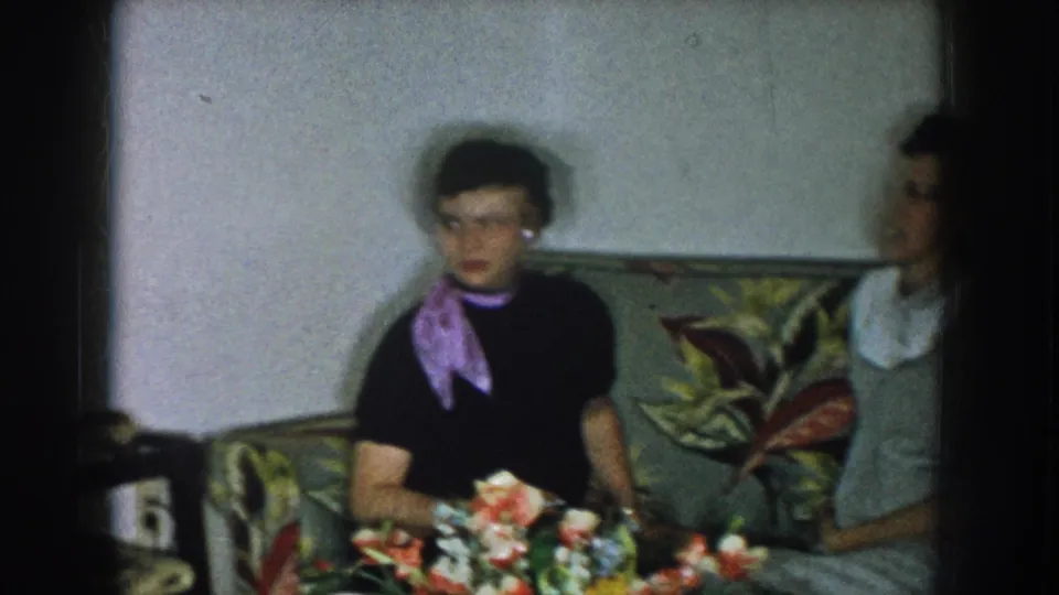 Two women in late-1950s daywear on a floral-upholstered sofa in a domestic living room; a colorful flower bouquet sits on a surface in the foreground.
