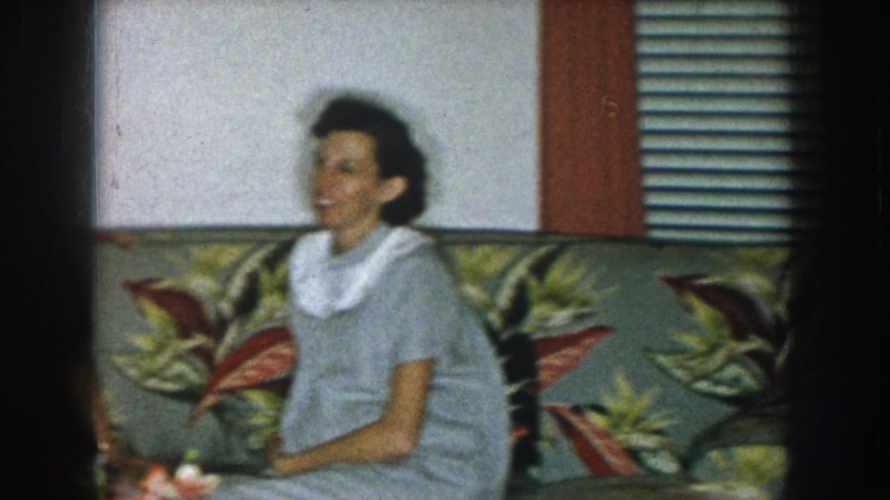 Single woman in a light gray dress with white collar seated on a floral sofa, smiling; venetian blinds and a red-framed window edge visible at right.