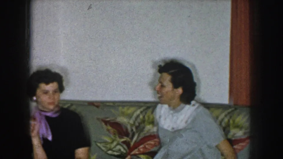 Two women in mid-century daywear on a tropical-print sofa in a domestic living room, in relaxed conversation during what appears to be a family gathering.