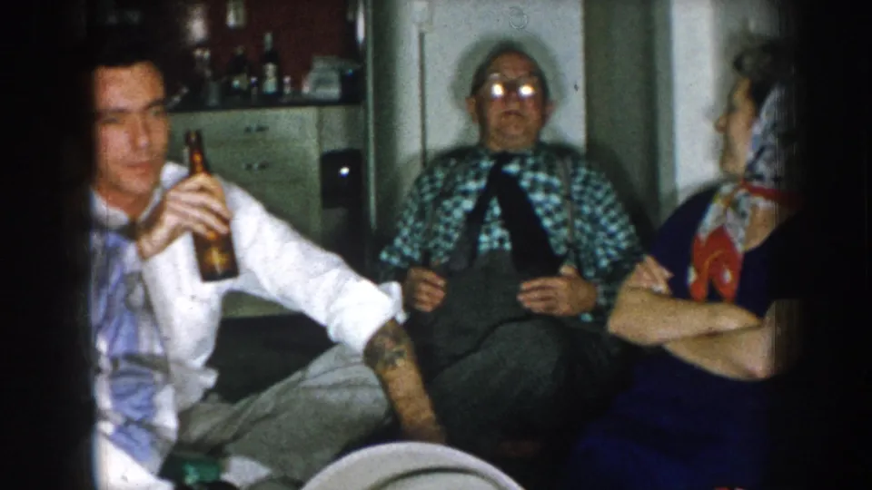 A young man holding a beer bottle in a domestic living room, with older adults in casual 1950s dress seated behind him; a shelf with bottles is visible in the background.