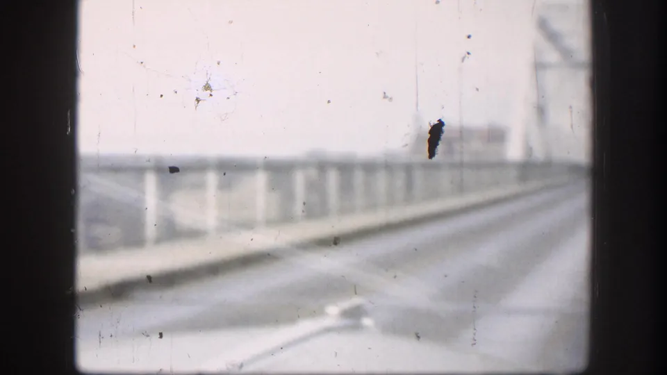 8mm home-movie shot through a car side window on a bridge crossing circa 1958, showing guardrails, distant bridge superstructure, overcast sky, and a blue film artifact.