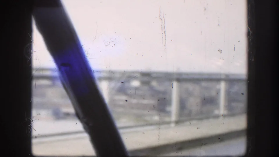 Final 8mm frame of a 1958 bridge crossing — a dark windshield wiper arm and vivid blue artifact dominate the foreground, with bridge guardrails and hazy shoreline behind.