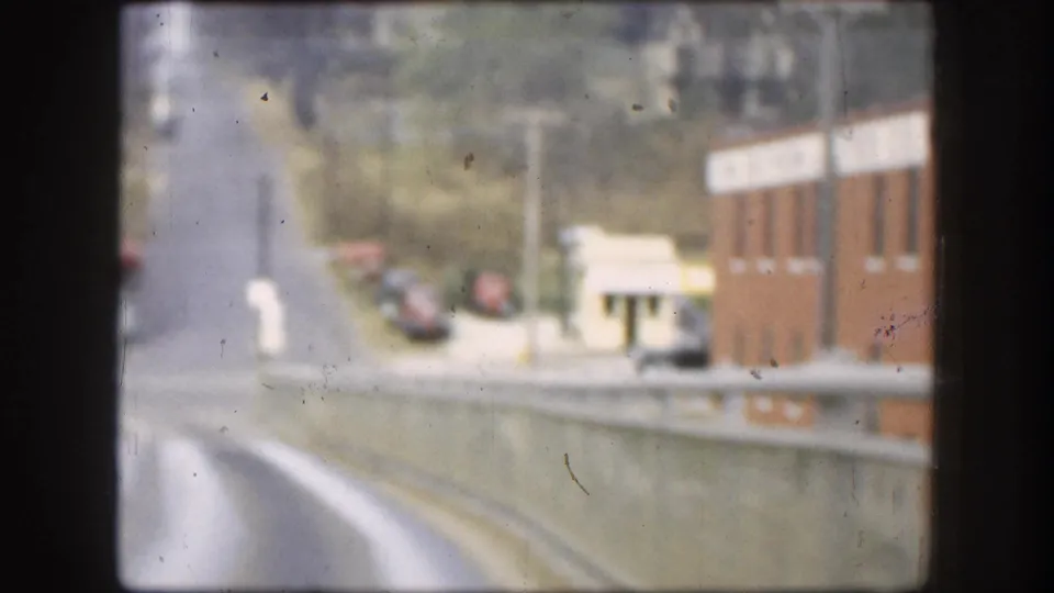 8mm frame from an elevated roadway looking down at a 1950s street scene with a brick building, vintage parked cars, and utility poles.