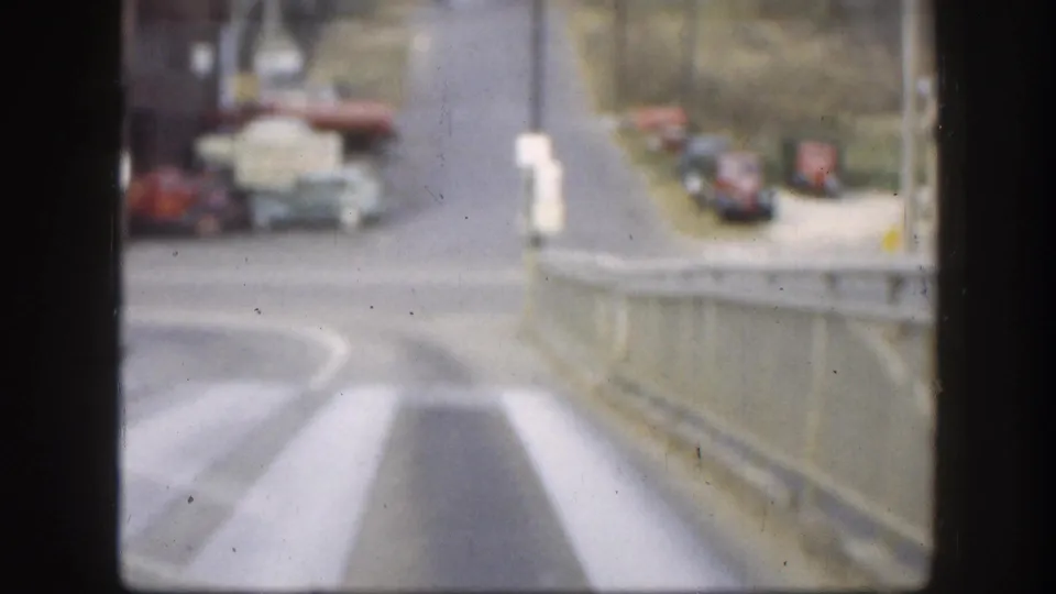 8mm frame showing a 1950s American highway with vintage trucks and cars, painted lane markings, and a white roadside marker.