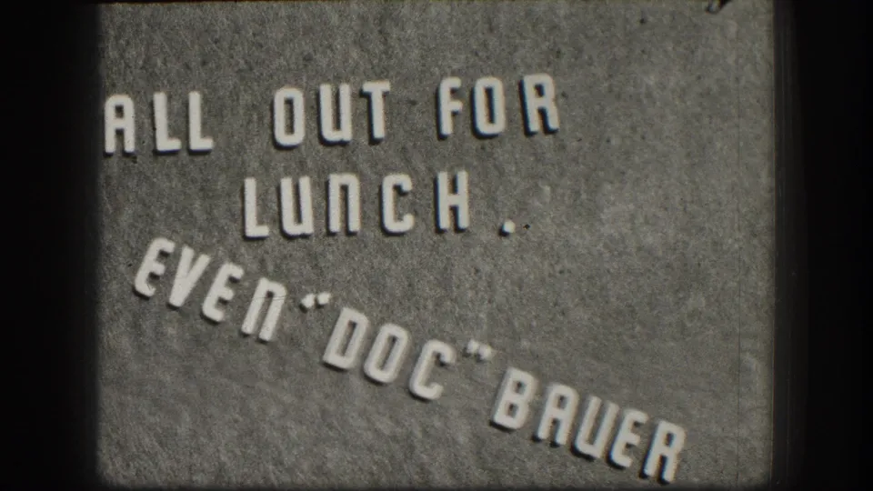 8mm home movie intertitle circa 1958 with embossed-style text reading 'ALL OUT FOR LUNCH. EVEN DOC BAUER' on a gray textured background.