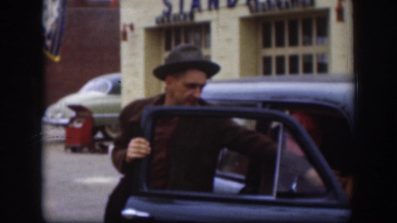 Man in dark fedora and brown jacket reaching for the open door of a dark classic American sedan on a commercial street.