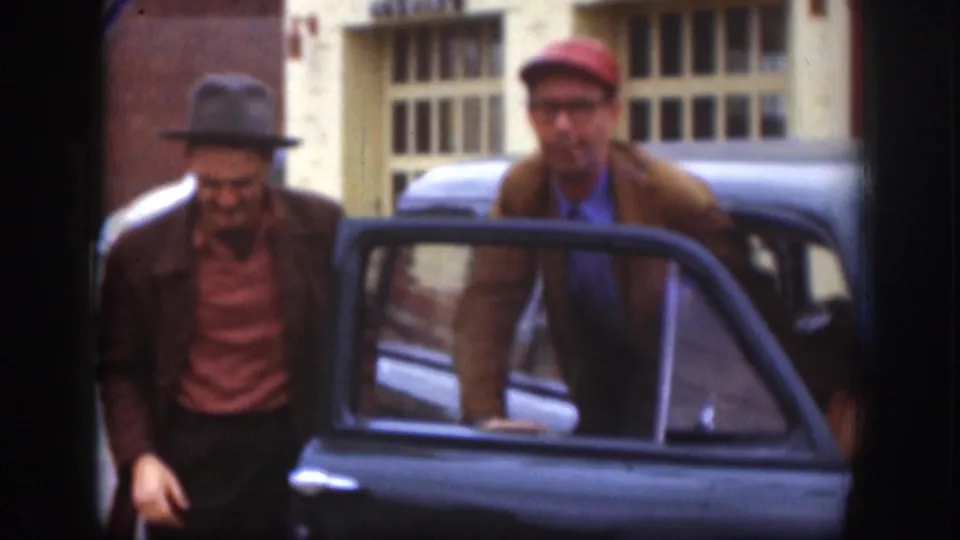 Two men — one in a gray fedora and striped shirt, one in a red cap and glasses — beside a dark classic sedan with its door open; brick building with gridded windows behind them, Kansas circa 1958.