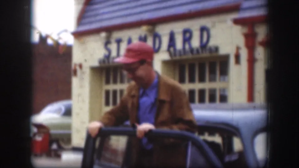 Man in red cap and brown jacket beside a dark classic sedan; a building with a prominent blue 'STANDARD' sign and red trim occupies the background, Kansas circa 1958.