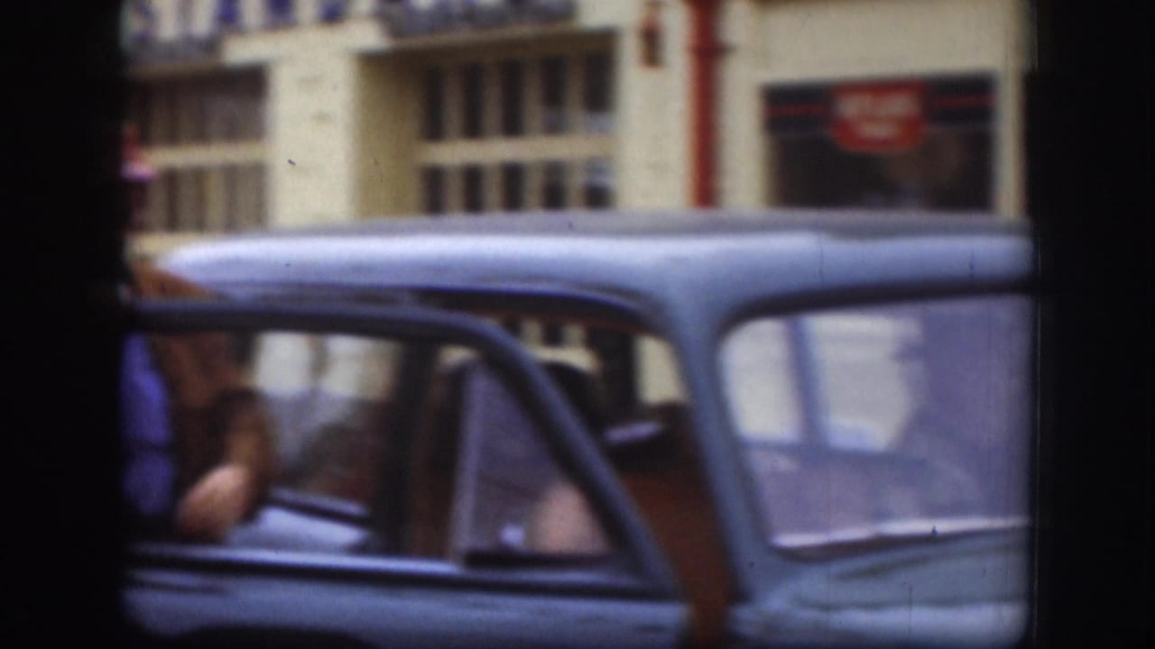 Close shot of a late-1950s dark American sedan in Kansas; a person partially visible at the open passenger door with an Atlas Tires storefront behind, circa 1958.