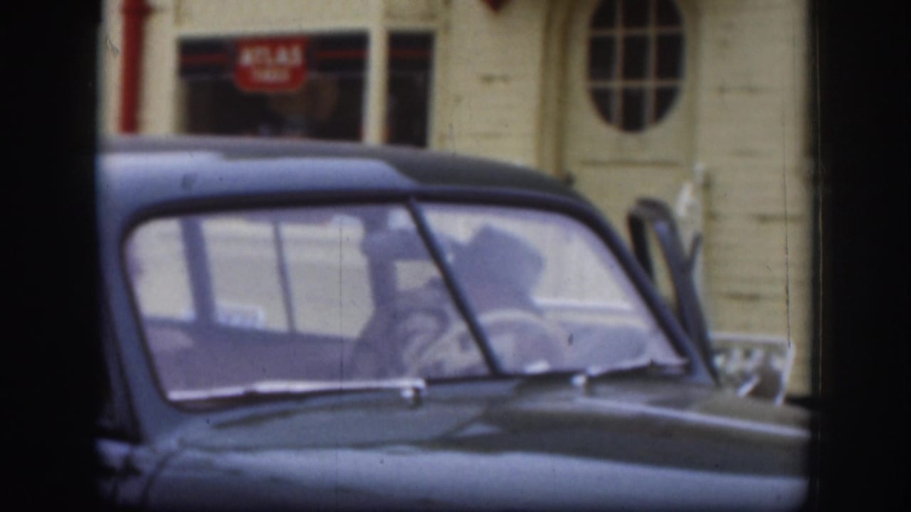 Dark American sedan with figure visible inside, cream and red building with Atlas Tires sign in background.