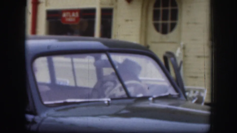 Late-1950s dark American sedan with a figure visible through the window; Atlas Tires storefront and arched oval window on cream-coloured building behind, Kansas, circa 1958.