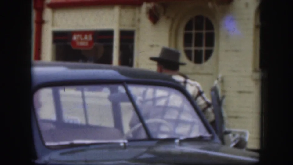 Man in a brimmed hat stands alongside a 1950s dark-coloured sedan on a Kansas street; an Atlas Tires storefront and arched oval window are visible on the cream building behind, circa 1958.