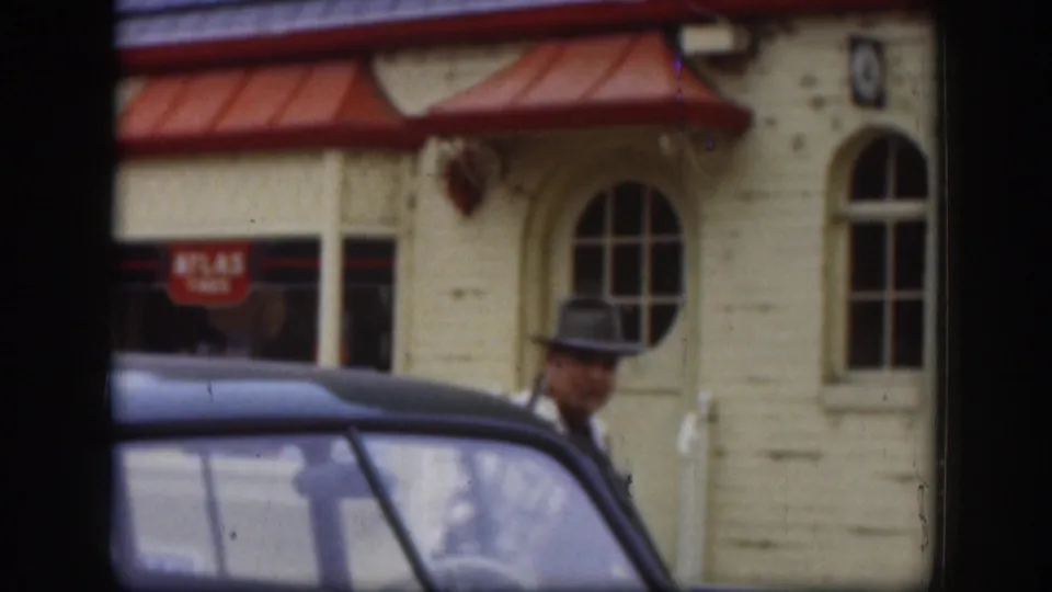 Man in a brimmed hat stands past a 1950s sedan; a red-awninged commercial building with Atlas Tires signage and oval arched window is visible behind him, Kansas, circa 1958.