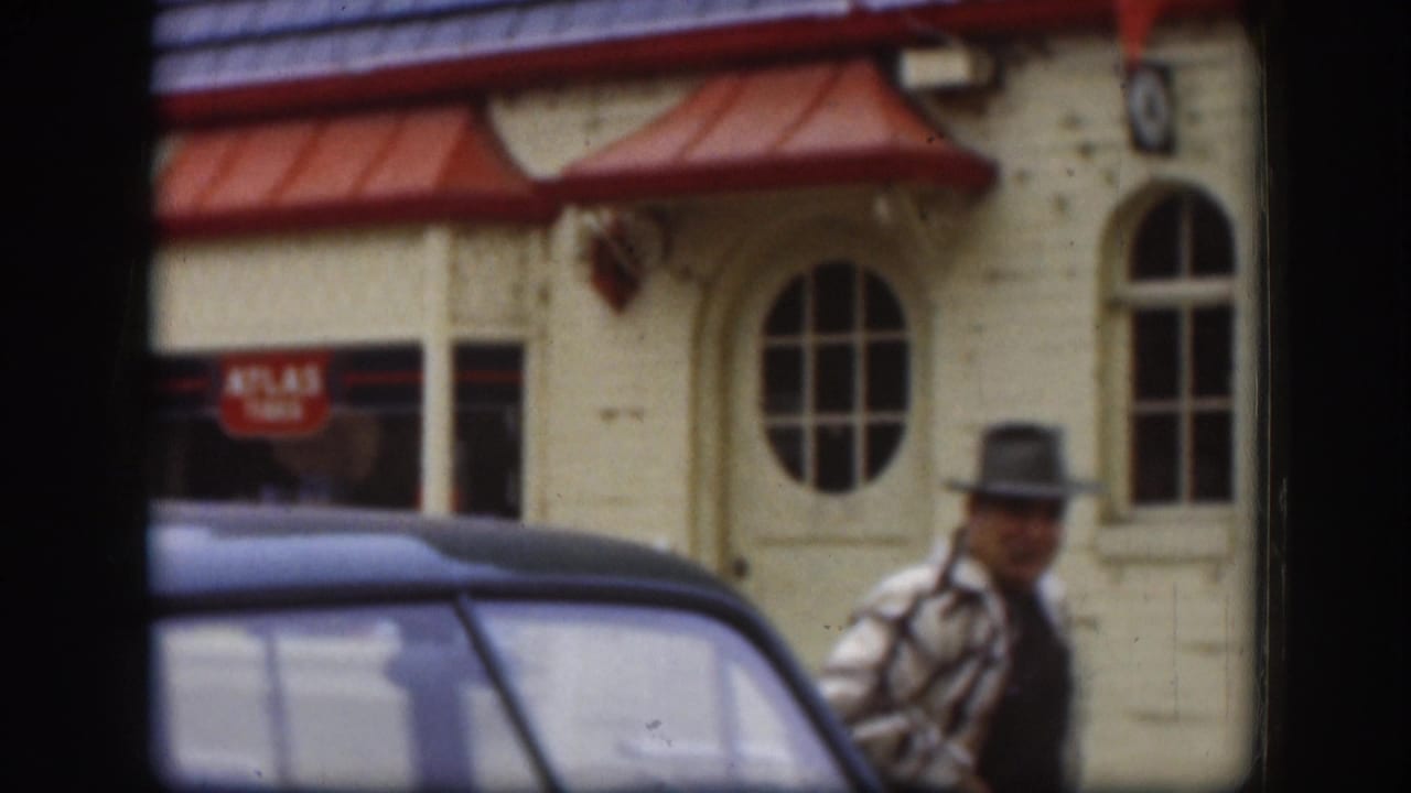 Man in dark fedora and plaid jacket passes a cream-painted commercial building with red awnings and ATLAS TIRES signage, blue classic car at left; Kansas, circa 1958.