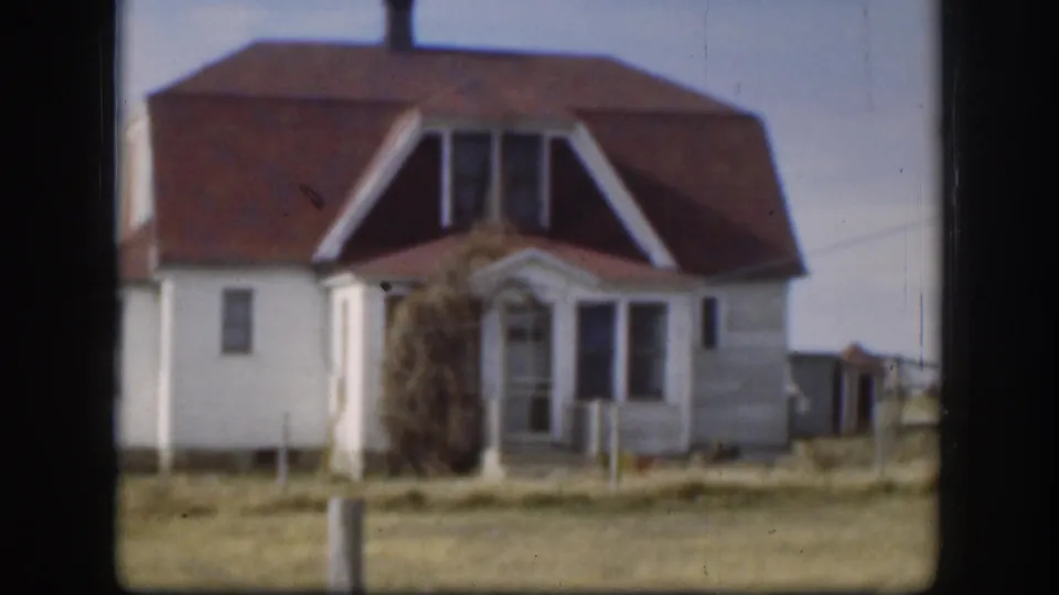 Exterior of a white clapboard farmhouse with a red-brown hip roof and dormer gables, flat open landscape behind, consistent with rural Kansas circa 1958.