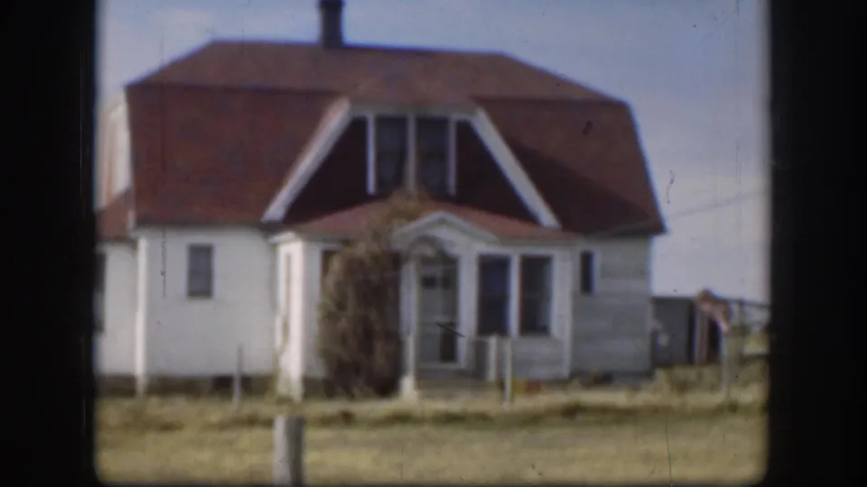 Continued exterior view of the same white clapboard farmhouse, red-brown roof and dormer peaks, flat Kansas landscape under pale sky, circa 1958.