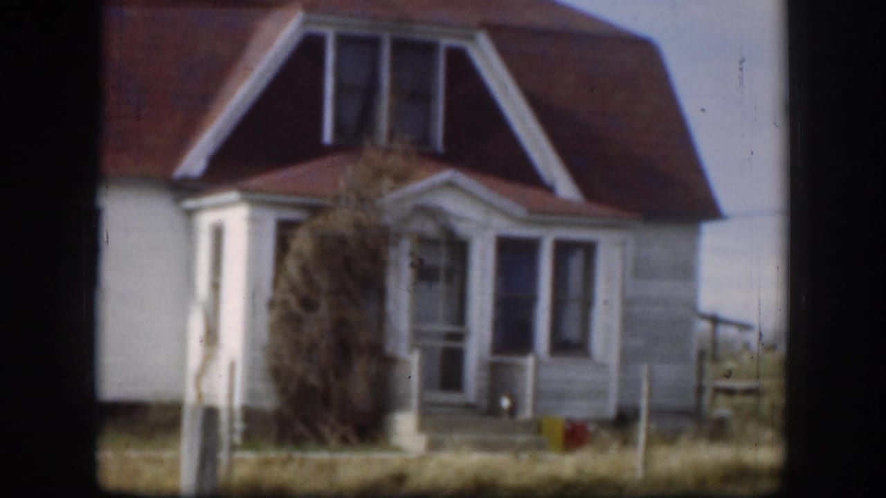 1958 Kansas farmhouse, closer angle: red gambrel roof, white dormer trim, large dried brush or tumbleweed mass obscuring porch, small red object at base.