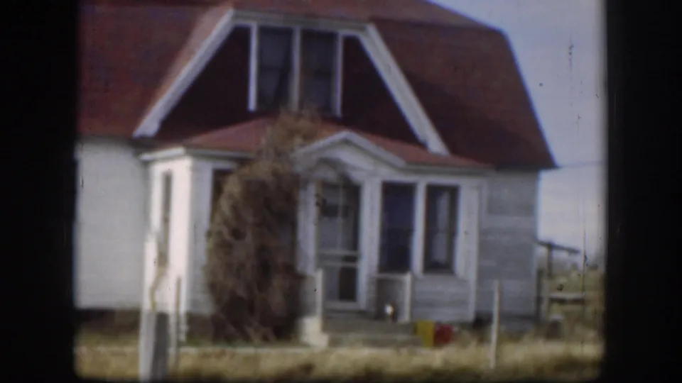 1958 Kansas farmhouse, closer angle: red gambrel roof, white dormer trim, large dried brush or tumbleweed mass obscuring porch, small red object at base.