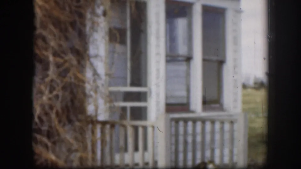 1958 Kansas farmhouse: tight zoom on tangled dried brush mass against white siding; white porch balusters visible behind, red roof at top edge.