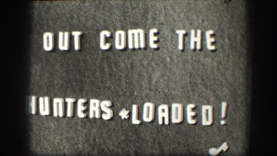 8mm home-movie title card circa 1958: 'OUT COME THE HUNTERS * LOADED!' in hand-set block letters on dark board, likely a chapter opener for a Kansas hunting-trip reel.