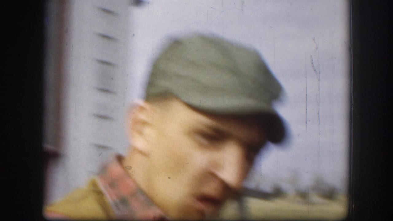 Close-up of a man in a green flat cap looking downward, wearing an olive hunting vest, with white clapboard siding visible behind him.