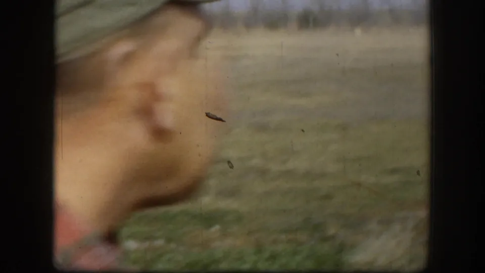 Motion-blurred 8mm frame: hand in foreground and flat open field behind — apparent camera movement or subject gesture mid-shot, consistent with amateur field filming.