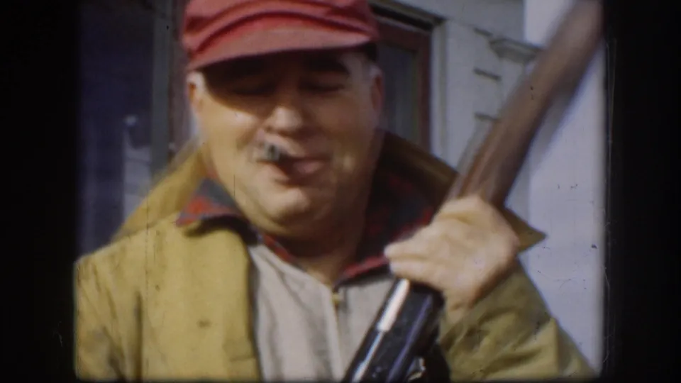 Hunter in red-billed cap grinning directly at camera, cigar in mouth, rifle shouldered with barrel raised, white clapboard siding behind — Kansas, circa 1958.