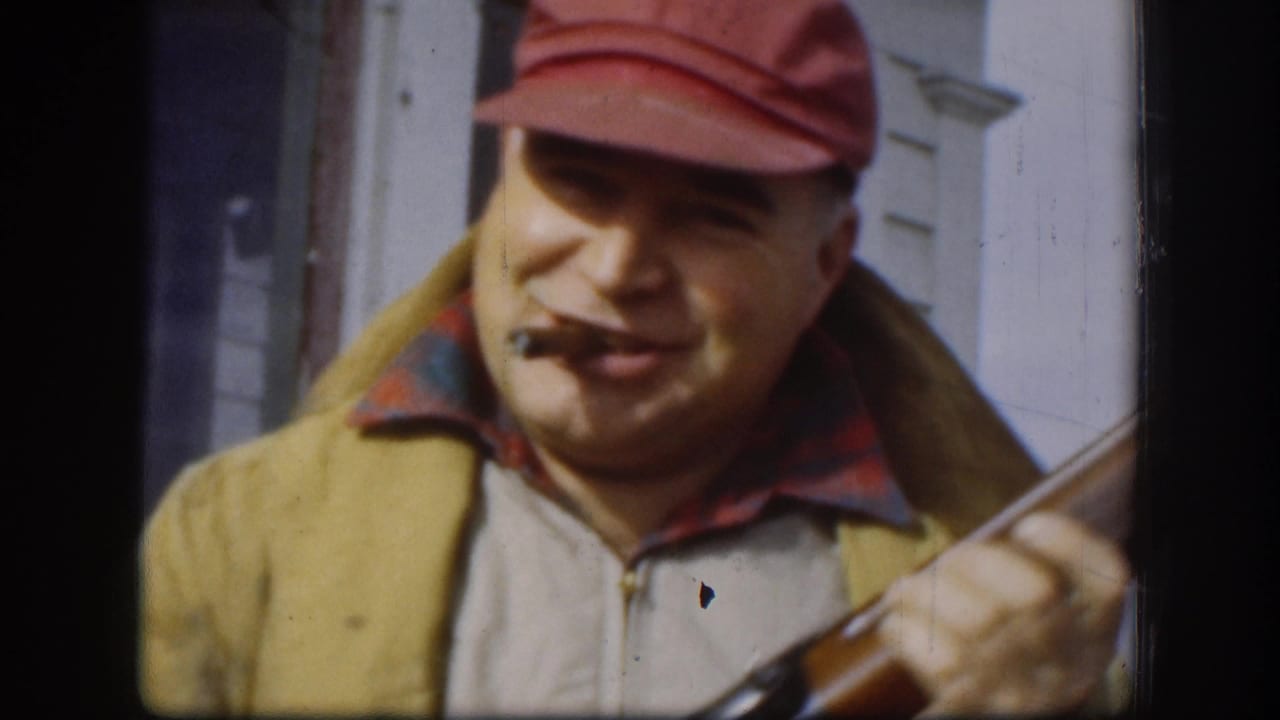 Middle-aged man in red bill cap and yellow zip jacket grips a rifle while smoking a cigar; white clapboard house siding behind him; Kansas, circa 1958.