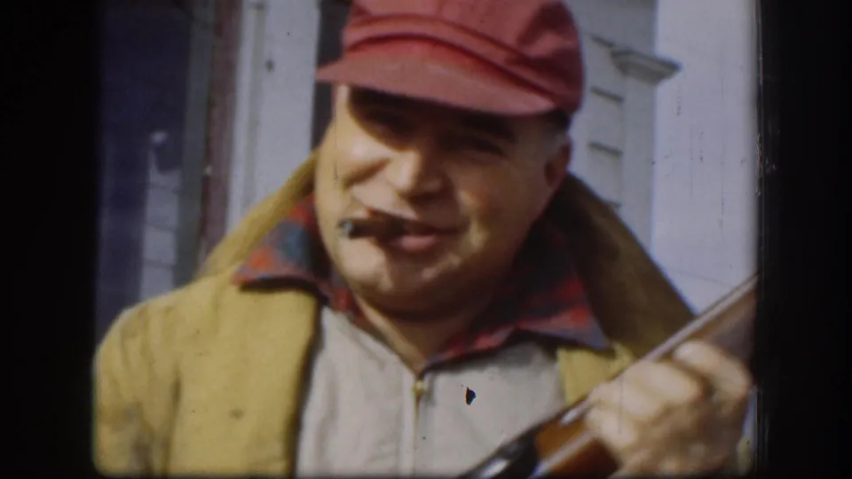 Middle-aged man in red bill cap and yellow zip jacket grips a rifle while smoking a cigar; white clapboard house siding behind him; Kansas, circa 1958.