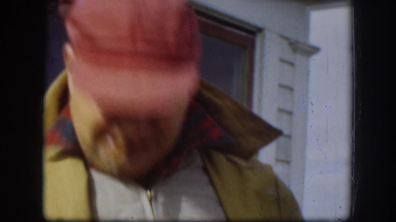 Tilted close frame showing top of man's red cap and yellow jacket; white house siding and window trim visible; face not shown.