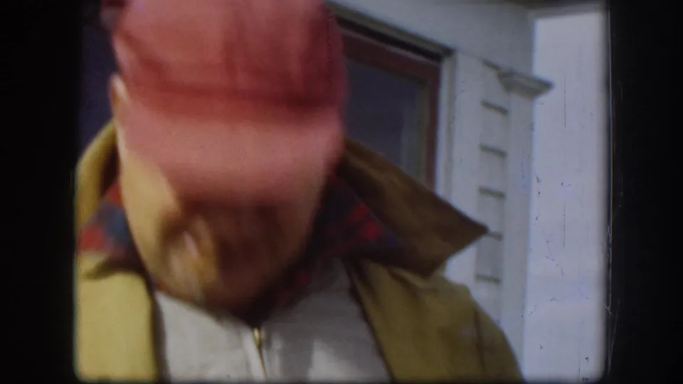 Tilted close frame showing the crown of a red bill cap and yellow zip jacket; white clapboard house exterior in background; handheld camera drift evident, Kansas, circa 1958.
