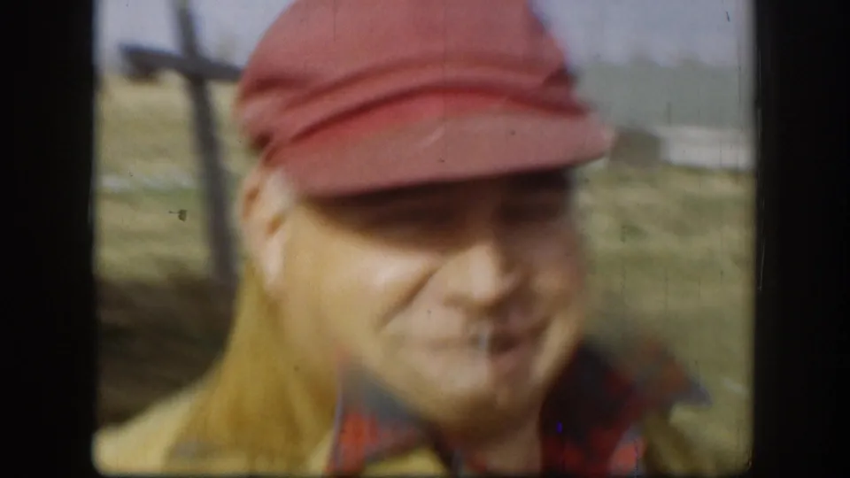 Near-focus close-up of a man in a red bill cap; green field and house wall behind him; 8mm handheld drift creates soft focus; Kansas, circa 1958.