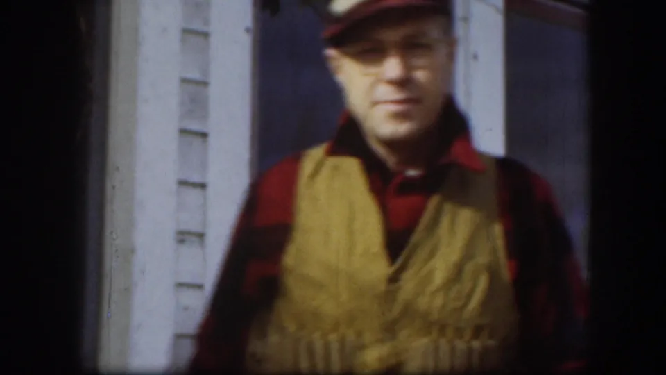 Man in red bill cap, red plaid shirt, and tan quilted vest poses in front of white clapboard house exterior with louvered shutters; Kansas, circa 1958.